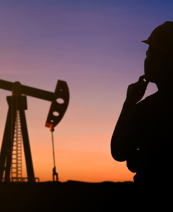 "Oil field engineer silhouette at sunset with pumpjack oil rig in background wearing safety helmet"